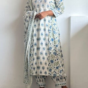 A woman wearing a soft blue Kalamkari three-piece set with navy and sage floral prints, featuring a V-neck tunic, ankle-length bottoms, and a lightweight dupatta. She pairs it with black sandals and poses against a white wall.