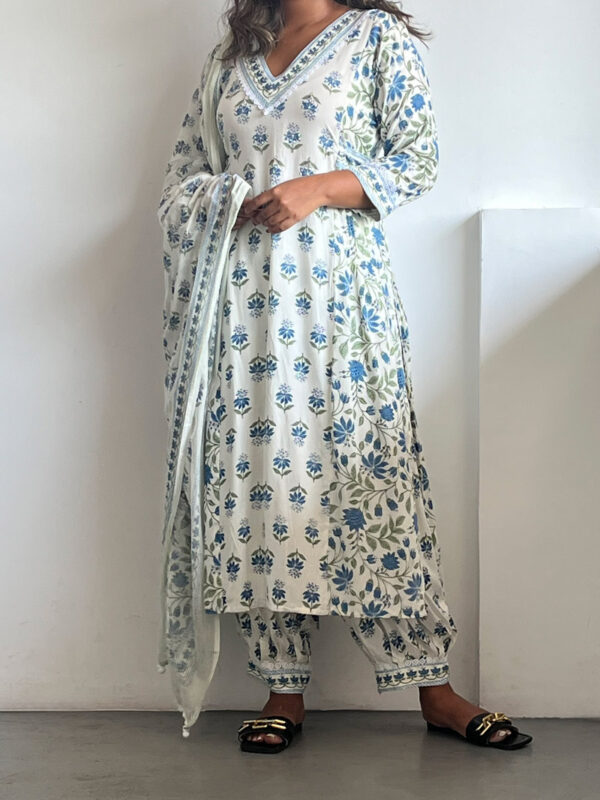 A woman wearing a soft blue Kalamkari three-piece set with navy and sage floral prints, featuring a V-neck tunic, ankle-length bottoms, and a lightweight dupatta. She pairs it with black sandals and poses against a white wall.