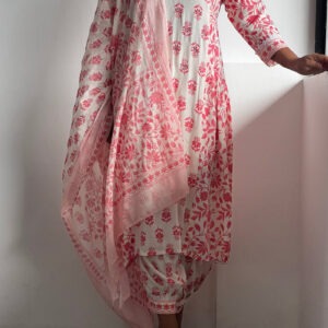 A woman wearing a blush pink Kalamkari set with a V-neck tunic, printed bottoms, and a lightweight dupatta, posing against a white wall.