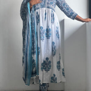 A woman wearing an ocean blue Kalamkari set with a gathered waist tunic, printed bottoms, and a tassel-detailed dupatta, posing against a white wall.