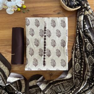 Unstitched 3-piece set with beige floral printed top, plain dark brown pant material, and a tribal printed dupatta with tassels laid flat on a wooden table.