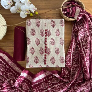 Unstitched 3-piece material set with beige floral printed top, plain wine pant fabric, and tribal-style dupatta with tassels laid on a wooden table.