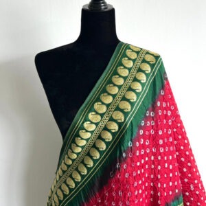 Red Bandhani dupatta draped on a black mannequin, featuring intricate white dotted motifs, green embroidered border with golden details, and a central paisley medallion. The corner tassels with ghungroo bells are partially visible.