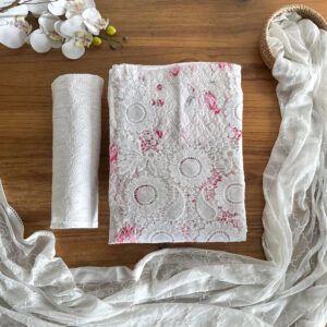 Angled flatlay of the white embroidered top with pink accents, paisley pant fabric, and sheer dupatta arranged on a wooden surface with orchid and basket decor.