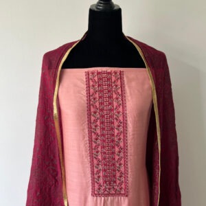 Close-up of a soft peach-pink unstitched material set displayed on a black mannequin. The top features detailed thread embroidery in deeper pink tones, while the matching dupatta in a rich maroon shade has gold and maroon woven patterns with a subtle sheen.