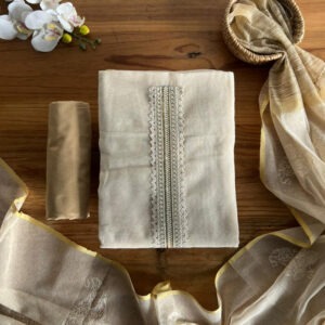 Ivory unstitched 3-piece set with lace-detailed top, beige pant, and golden dupatta laid on table.