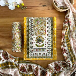 Mustard block print unstitched 3-piece material set laid on a flat table showing floral printed top, rolled pant material, and printed dupatta spread around.