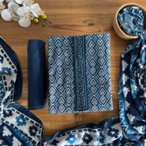 Deep indigo printed unstitched 3-piece material set laid flat on a wooden table, showcasing embroidered neckline detail, solid blue pant material, and a printed dupatta styled around the set.