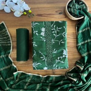 Unstitched emerald green three-piece suit set laid flat on a wooden surface, featuring a printed floral top material, solid pant fabric, and a sheer striped dupatta with gold lace edging.