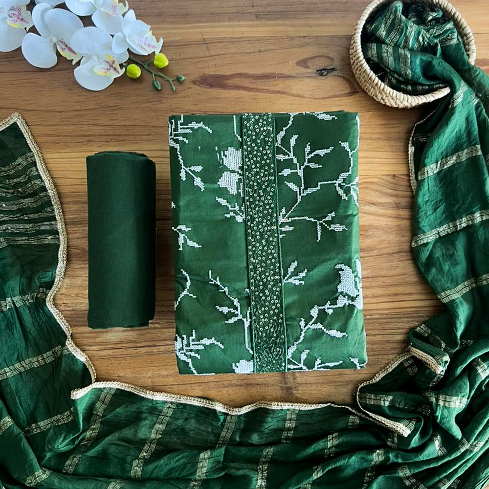 Unstitched emerald green three-piece suit set laid flat on a wooden surface, featuring a printed floral top material, solid pant fabric, and a sheer striped dupatta with gold lace edging.