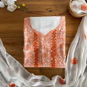 Ivory unstitched three-piece material set laid on a wooden table with orange floral embroidered top, sheer dupatta with matching orange motifs, and coordinated pant fabric