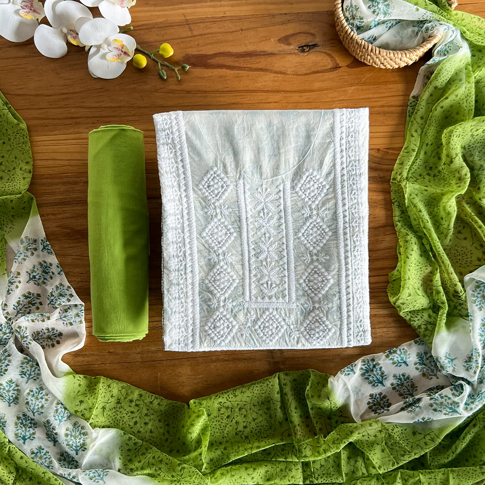 Pastel mint unstitched three-piece material set laid flat on a wooden table, featuring a white embroidered top, solid green pant fabric, and a lightweight printed dupatta arranged around the set